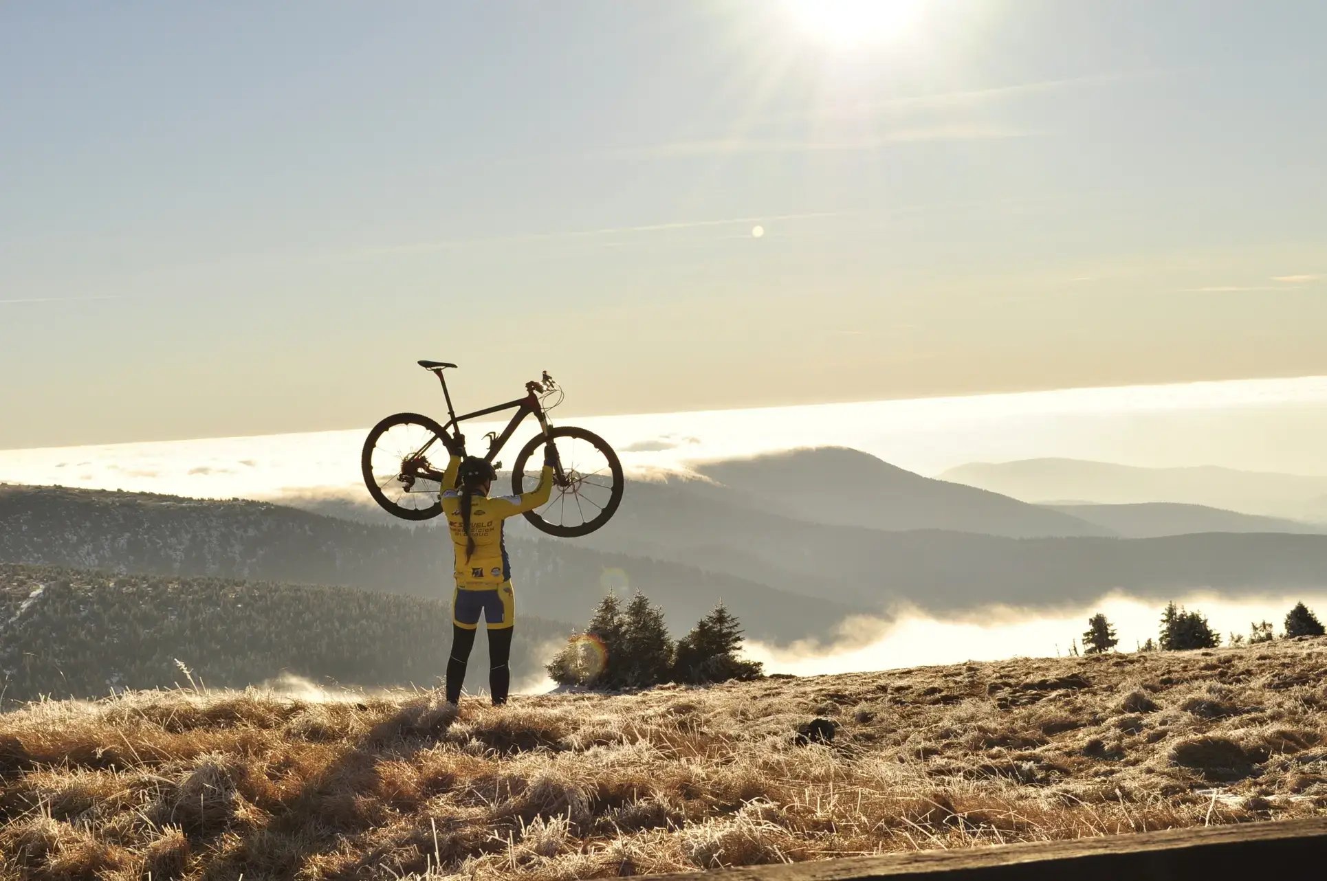 Cyclist in yellow gear triumphantly holding a bicycle overhead, standing on a frosty hilltop with misty mountain peaks in the background. 