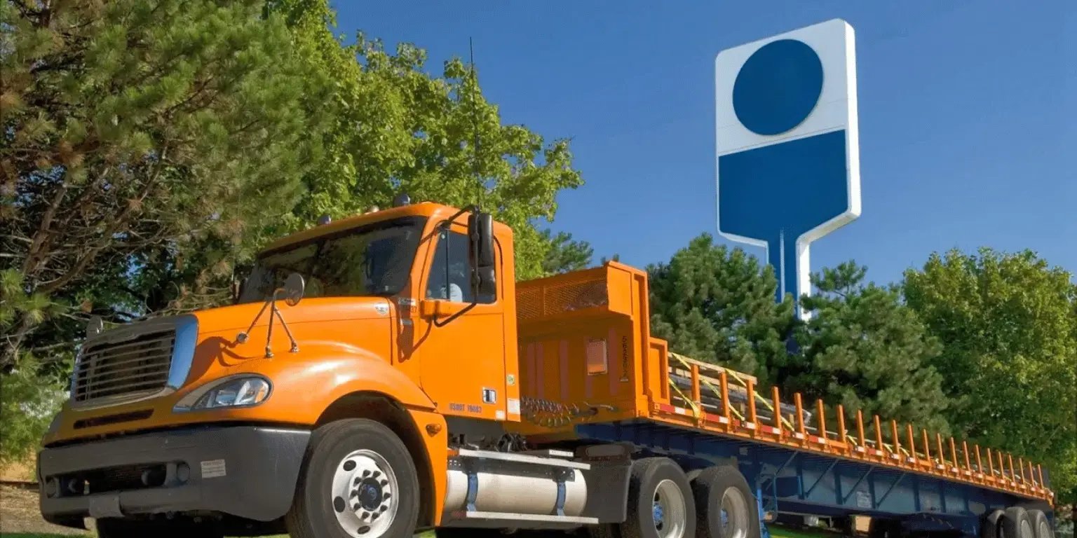  Bright orange flatbed truck parked outdoors on a sunny day, with green trees and a blue industrial building in the background. Ideal for heavy transport. 
