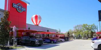 A row of cars parked under red shade canopies outside a building with a large red and white hot air balloon structure on the roof. The scene is set on a sunny day with a clear blue sky, suggesting an outdoor event or promotional display