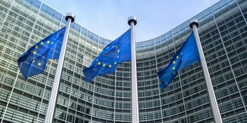 European Union flags flying in front of a modern glass building with a curved facade, representing an official EU institution or headquarters. The flags are blue with yellow stars, symbolizing unity among member states.