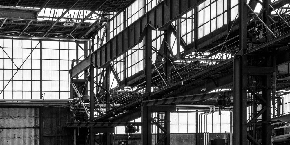 Black and white industrial interior with steel beams, large windows, and crisscrossing metal structures, highlighting a factory or warehouse setting. 