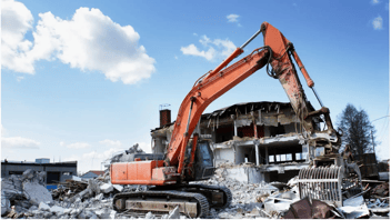 Excavator demolishing a building with rubble in the foreground and clear blue sky in the background.