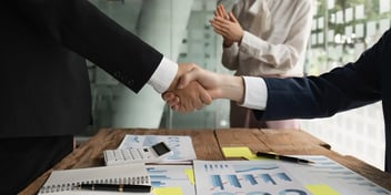 Two people shaking hands over a wooden table covered with business documents, charts, sticky notes, and a calculator, symbolizing a business agreement or partnership
