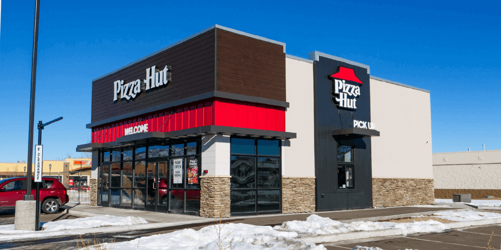  Exterior of a Pizza Hut restaurant with the recognizable red and black branding, featuring large windows and a modern design. Snow is visible on the ground, suggesting a cold or winter season setting. 