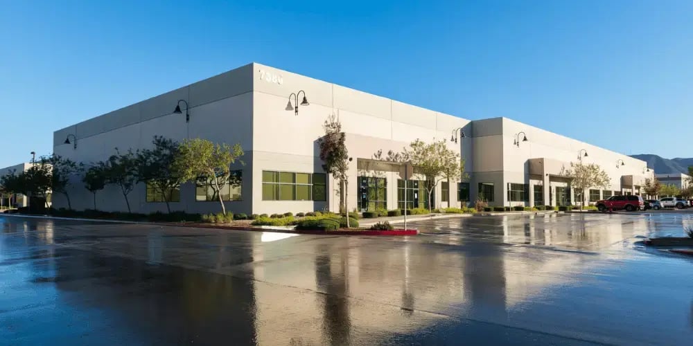  Modern industrial building with light-colored walls, large windows, and exterior lighting fixtures. Trees and landscaping surround the facility, with a clear blue sky above. 