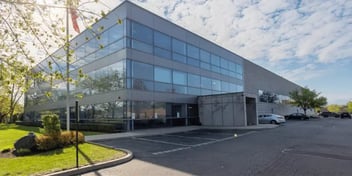 Exterior of the Widex USA building, a modern, multi-story office structure with large glass windows and a minimalistic design. The surrounding area includes a parking lot and landscaping, with trees visible in the background under a partly cloudy sky.