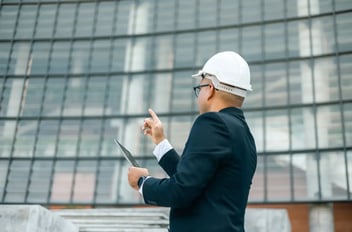 A businessman in a suit and hard hat gestures while holding a tablet near a glass building.