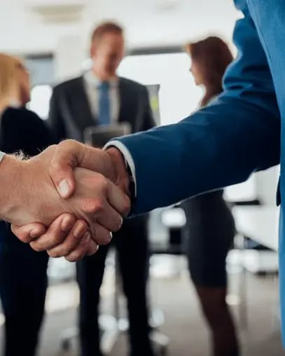 Two professionals shaking hands in a business setting, symbolizing partnership and collaboration, with blurred colleagues in the background.