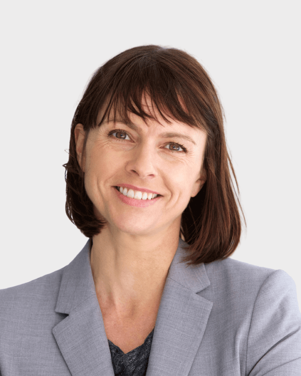 A professional woman with shoulder-length brown hair and bangs, smiling warmly while wearing a light gray blazer. She appears confident and approachable, set against a neutral background.