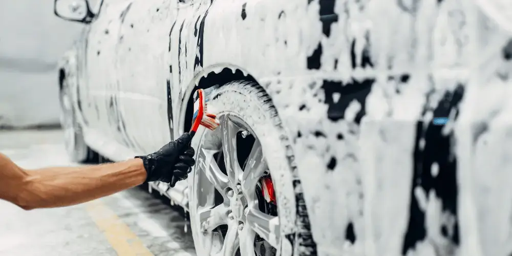 A close-up of a car wheel being cleaned with a soapy sponge, covered in thick white suds. A gloved hand is visible scrubbing the wheel, emphasizing attention to detail in the cleaning process