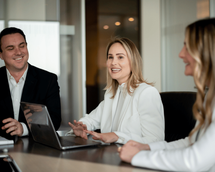 Nina, Chelsea, and Jack seated around a conference table, engaged in a lively discussion. Chelsea, in the center, is speaking with a smile while using a laptop, creating a collaborative and positive atmosphere. The setting suggests a professional meeting in a modern office environment.