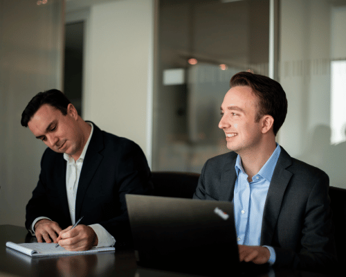 A close up photo of Spencer Freid and Jack Foley working at a desk together while jack writes on a paper and Spencer is looking up off screen across the table.